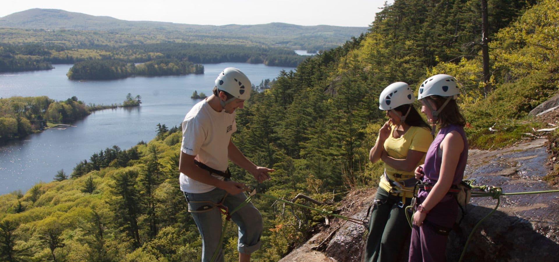 Three climbers in helmets discuss climbing techniques near a scenic lake surrounded by greenery.