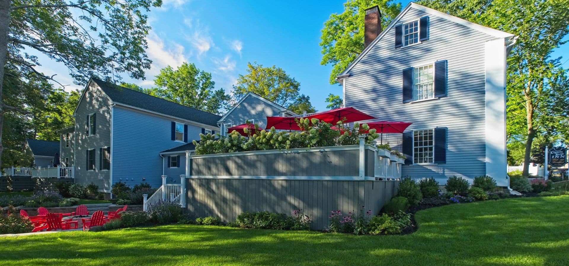 A blue house with a shaded deck, surrounded by lush greenery and red patio furniture.