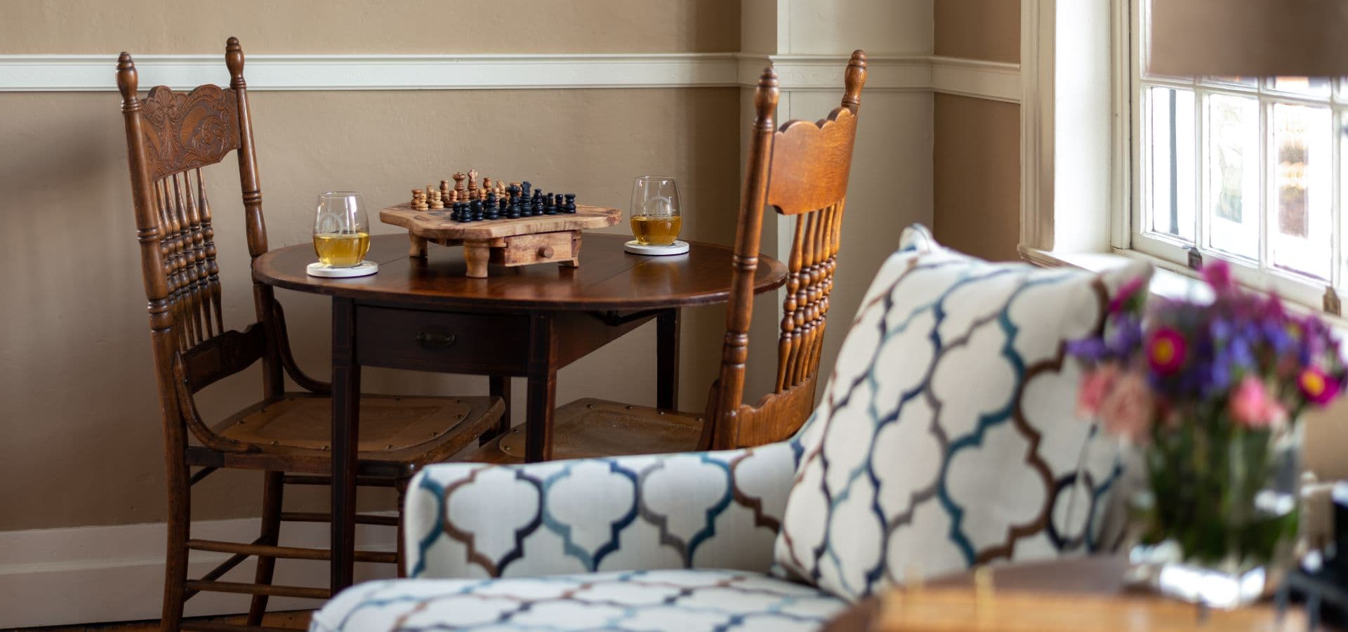 A cozy seating area features a small table with a chess set and two glasses, complemented by a patterned chair and a vase of flowers.