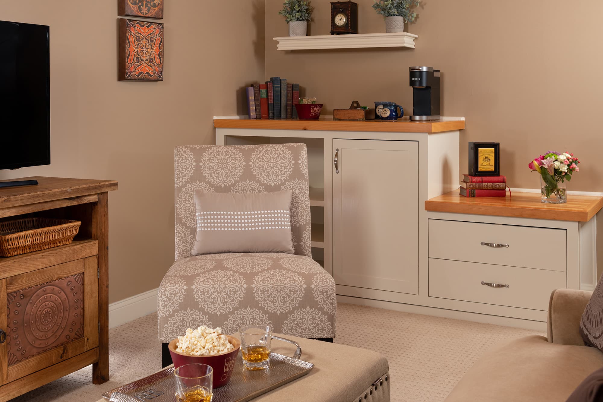 A cozy corner of a guest suite featuring a patterned slipper chair, a refreshment center with a Keurig coffee maker, and a wooden console. A tray with snacks and drinks sits in the foreground.
