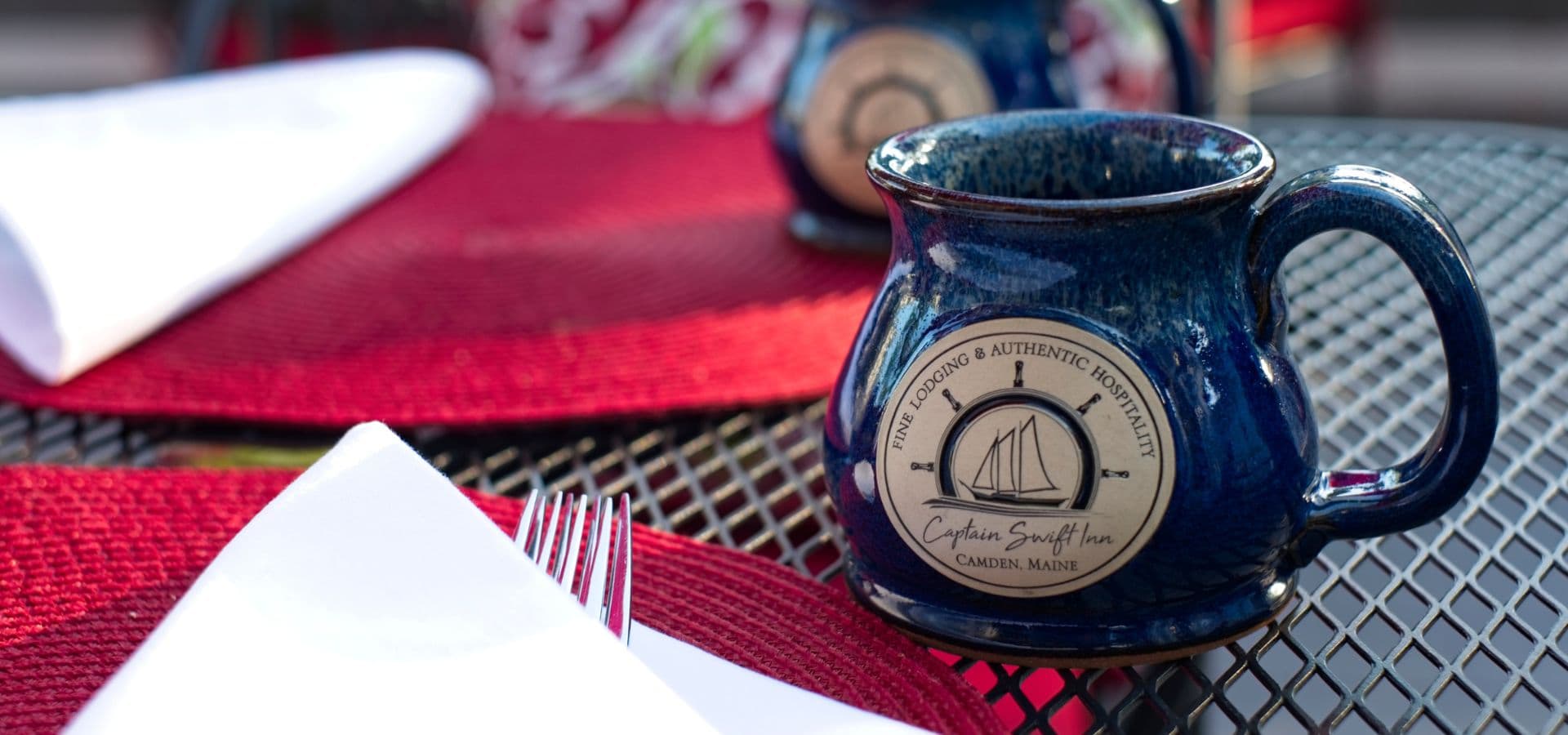A blue ceramic mug with a nautical logo sits on a table set with red placemats and folded napkins.