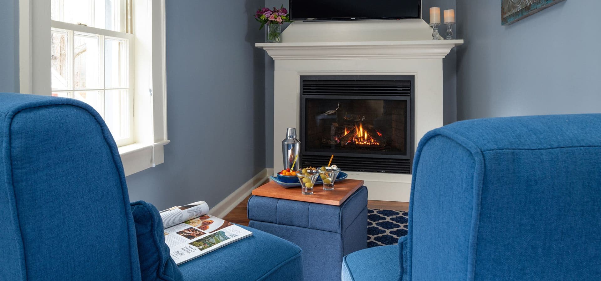 Cozy living space with blue chairs, a fireplace, and a small table displaying drinks and snacks.