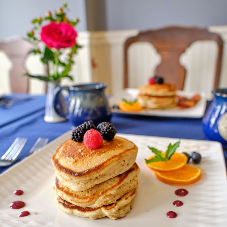 A plate of stacked pancakes topped with fresh berries, accompanied by orange slices and a decorative sauce, set on a table with a vase of flowers.