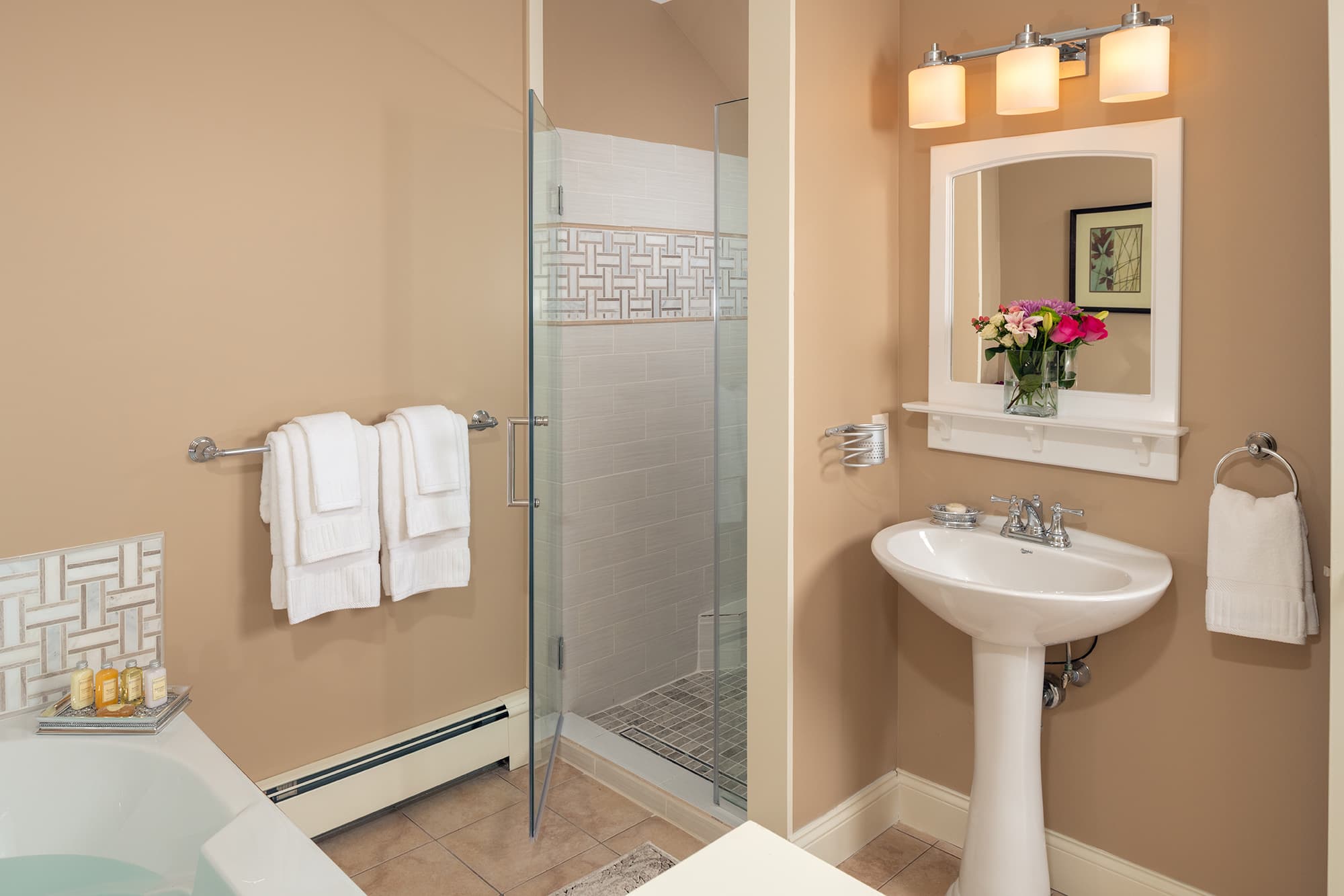 A warm-toned en-suite bathroom featuring a white pedestal sink, a glass-enclosed walk-in shower with decorative mosaic tiling, and a partial view of a whirlpool tub against tan walls.