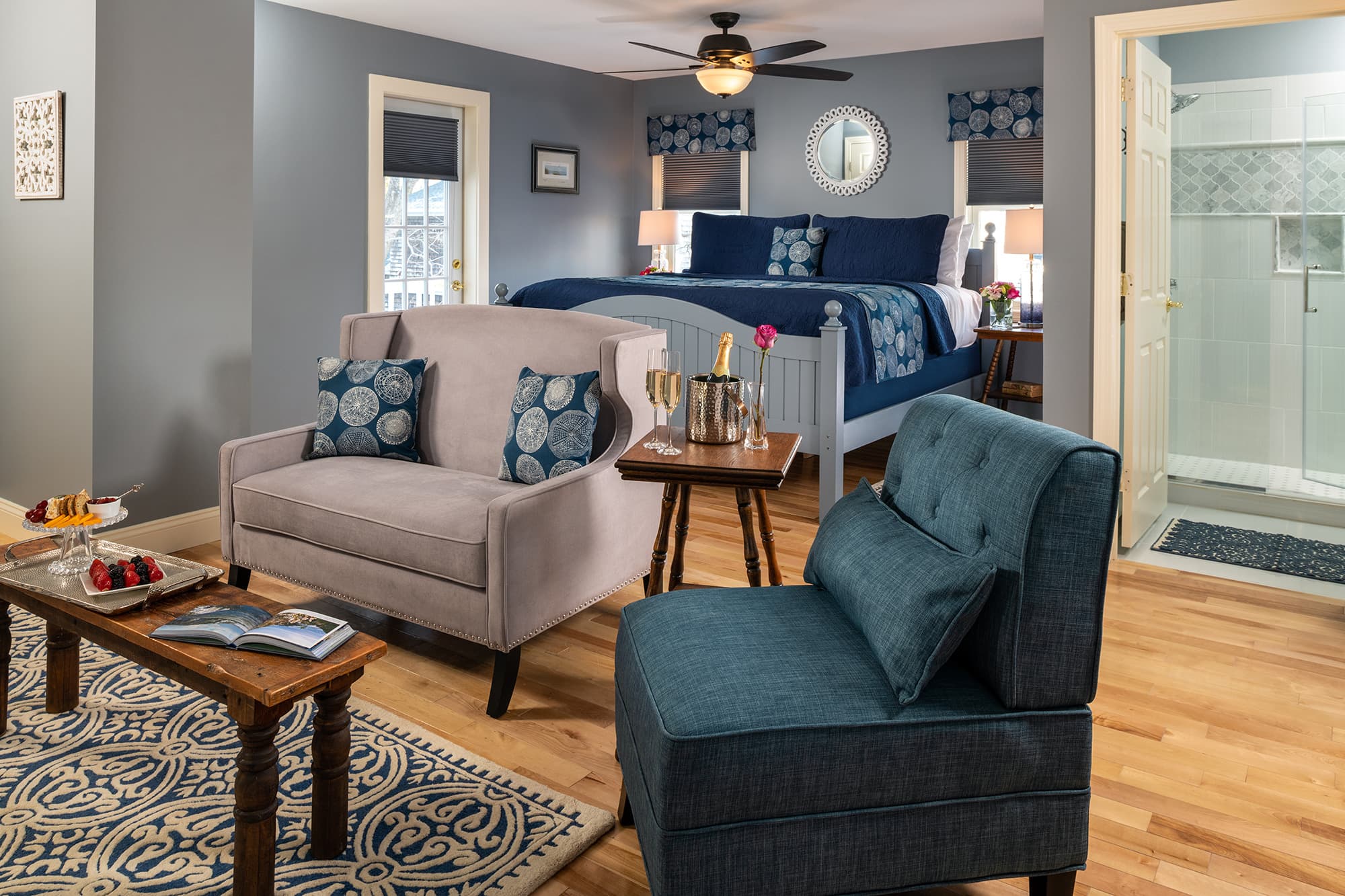 A spacious, modern guest suite with slate blue walls and hardwood floors, featuring a king bed with navy linens, a cozy seating area with a loveseat and teal armchair, and a view into a bright en-suite bathroom with a glass shower.