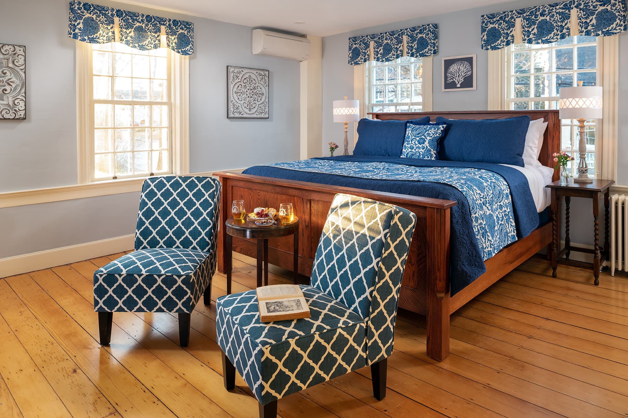 A sun-filled guestroom with light blue walls and wide-plank wood floors, featuring a wooden king-size sleigh bed with navy and patterned blue linens, two teal diamond-patterned chairs, and three large windows with decorative valances.
