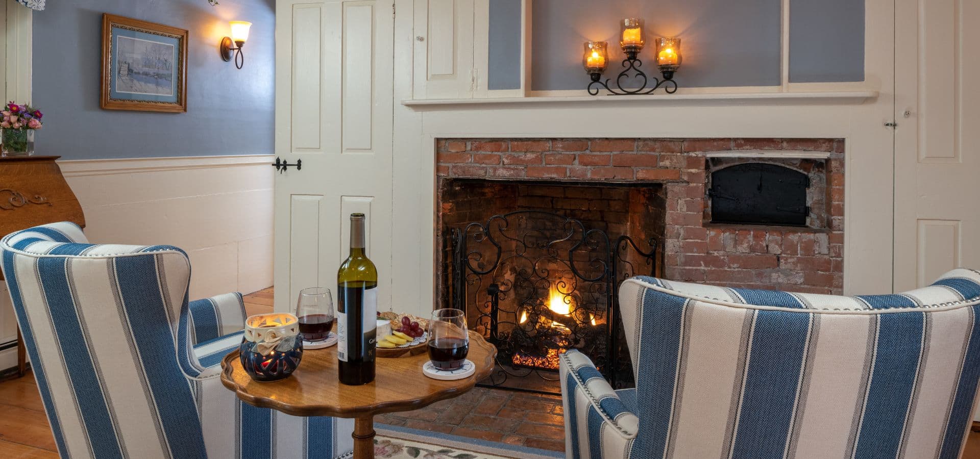 Cozy living room with a fireplace, two striped chairs, a small table with wine and snacks, and soft lighting.