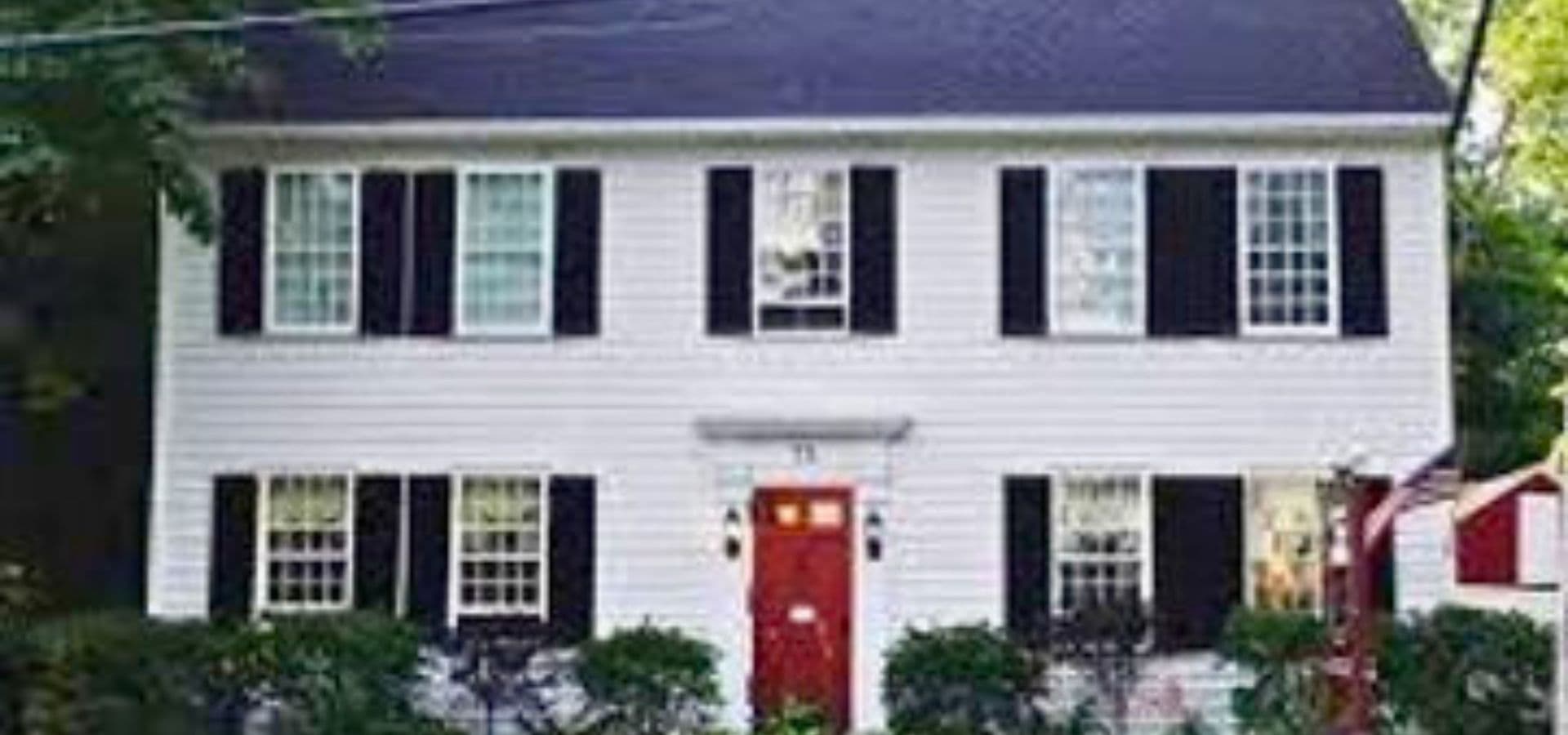 A two-story white house with black shutters and a red front door surrounded by greenery.