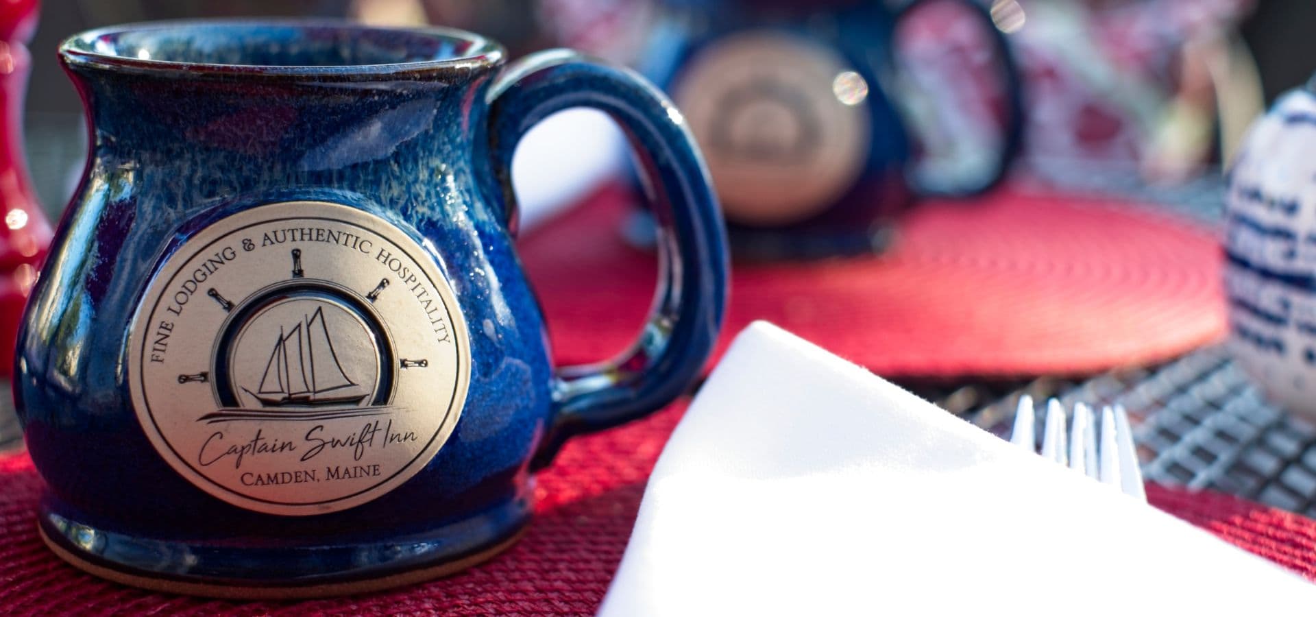 A blue ceramic mug with a logo from Captain Swift Inn in Camden, Maine, is placed on a red textured table setting.