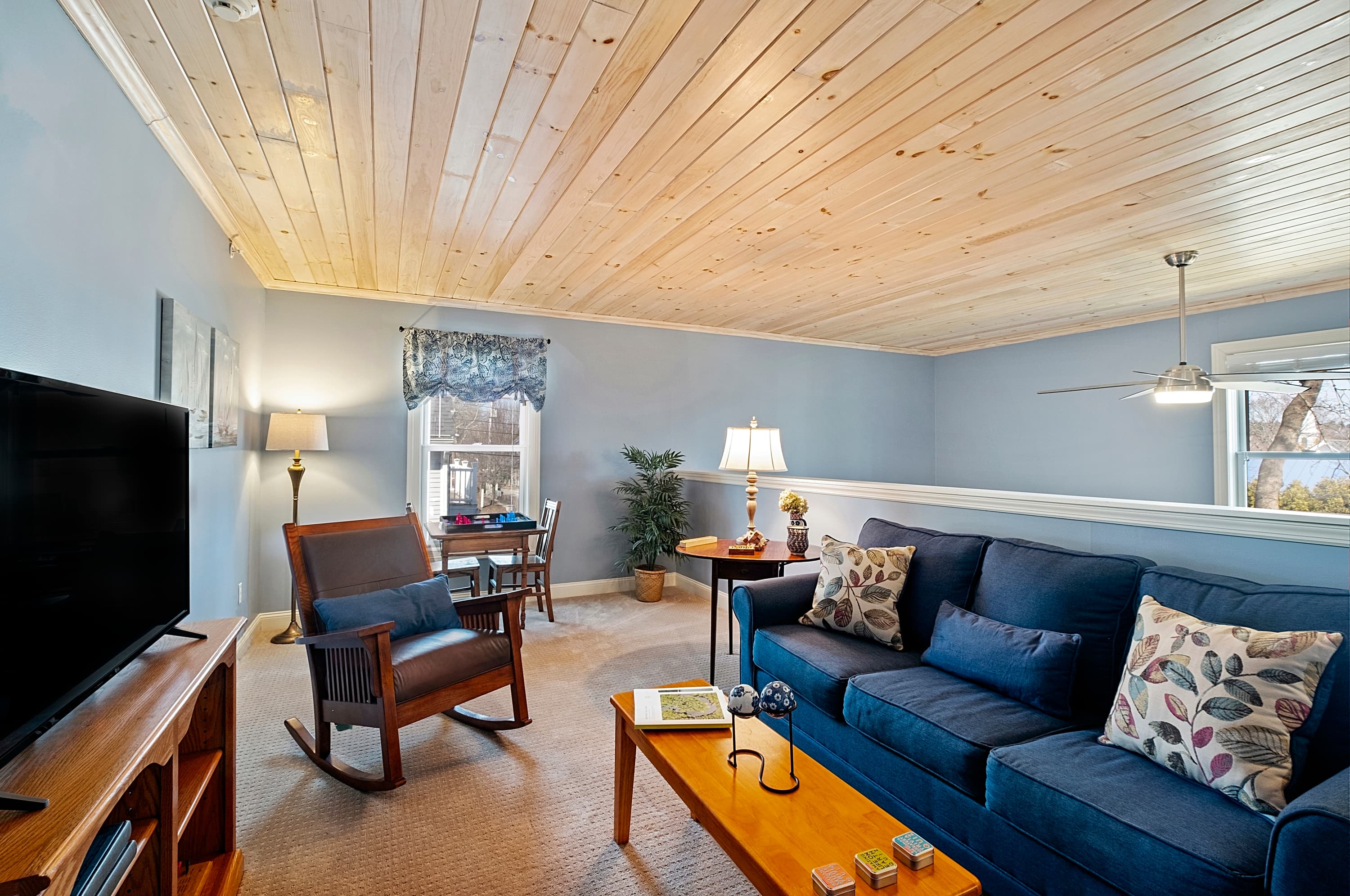 A cozy loft sitting area featuring a blue sofa, a wooden rocking chair, and a flat-screen TV, all under a light-colored wood plank ceiling with a small game table in the corner.