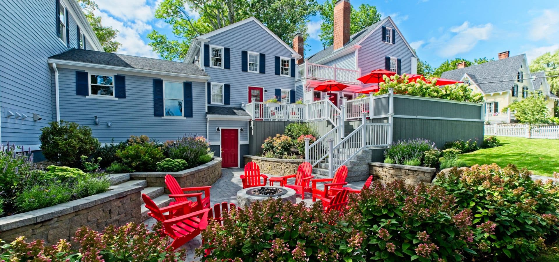 A backyard patio featuring red chairs, a fire pit, and houses surrounded by greenery under a blue sky.