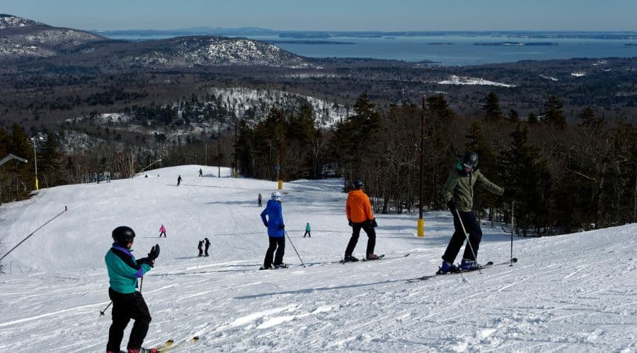 Skiers navigate a snowy slope with a scenic view of mountains and a lake in the background.