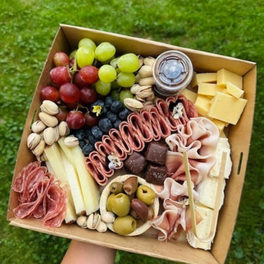 A gourmet charcuterie box featuring assorted cheeses, meats, fruits, nuts, and a small container of dip.