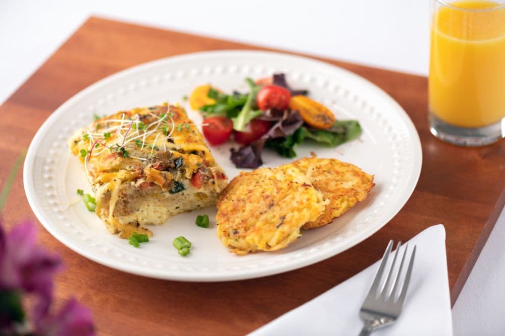 A plate with a vegetable frittata, two potato pancakes, a mixed salad, and a glass of orange juice.