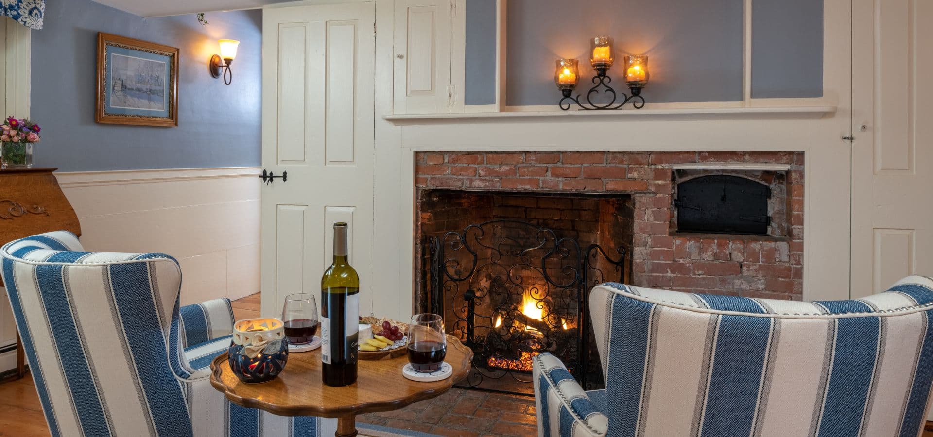 A cozy living room features two striped chairs, a small table with wine and snacks, and a warm fire in a brick fireplace.