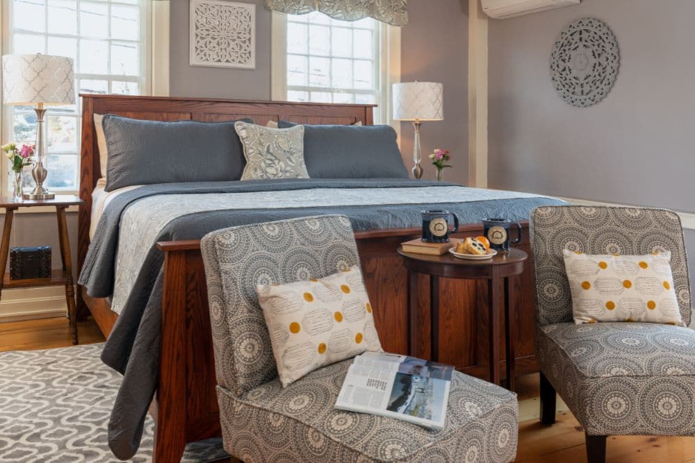 A cozy bedroom featuring a wooden bed with gray bedding, two patterned armchairs, and a small table with mugs and snacks.