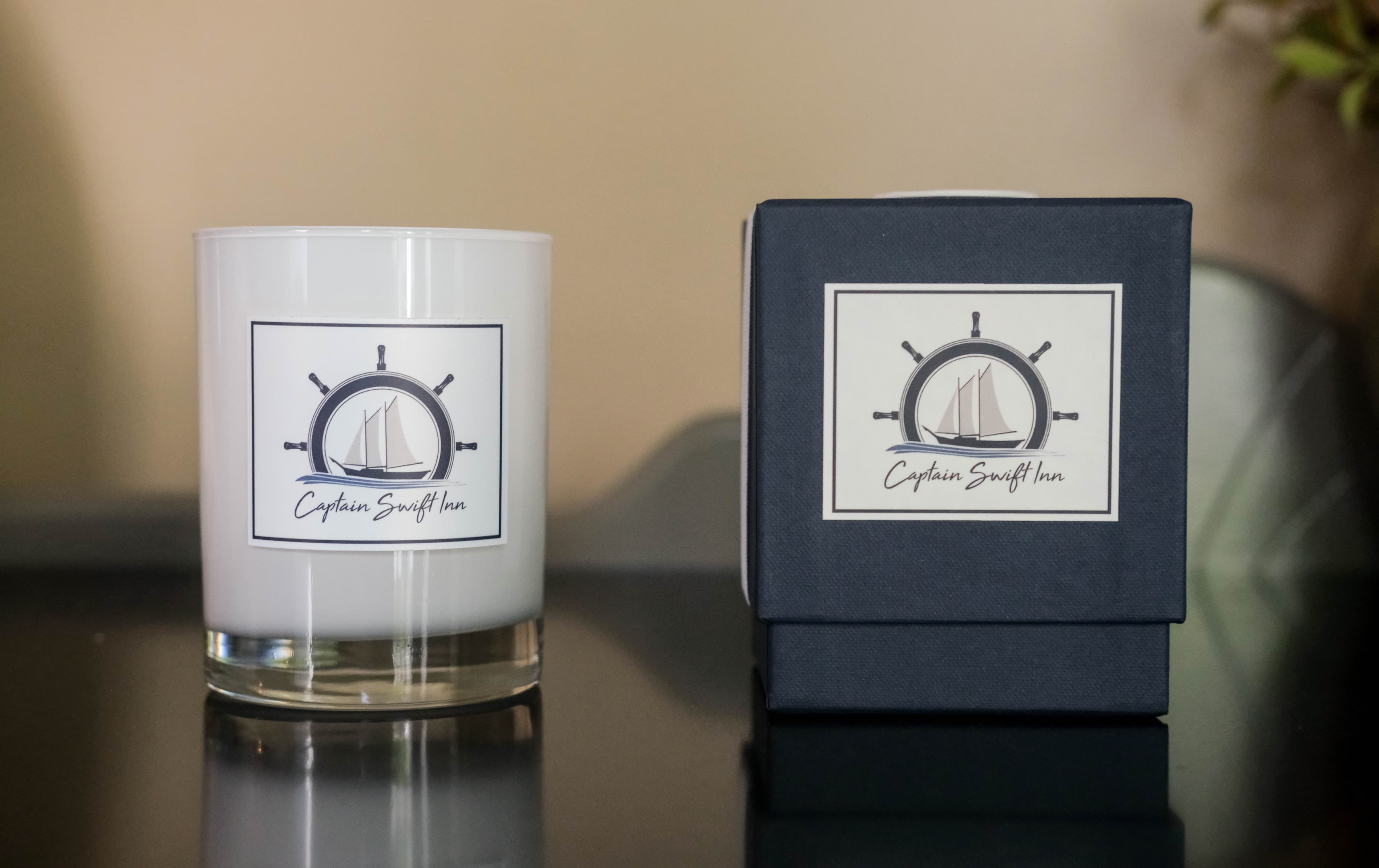 A candle with a logo featuring a ship's wheel and a sailboat, alongside its navy-blue box labeled "Captain Swift Inn."
