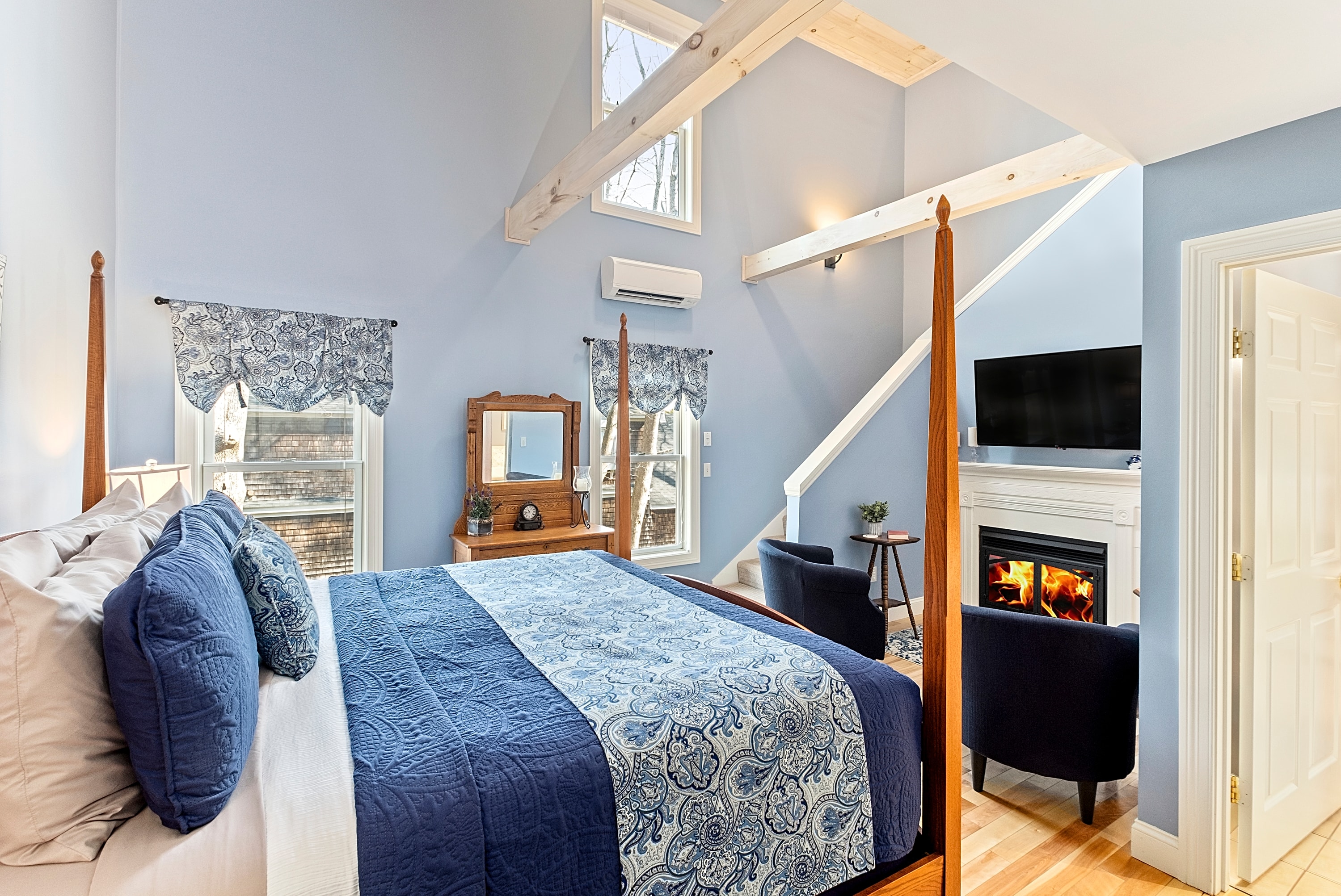 A bright, two-level luxury suite featuring a four-poster bed with blue linens, soaring ceilings with white beams, a gas fireplace, and a seating area with two blue velvet chairs.