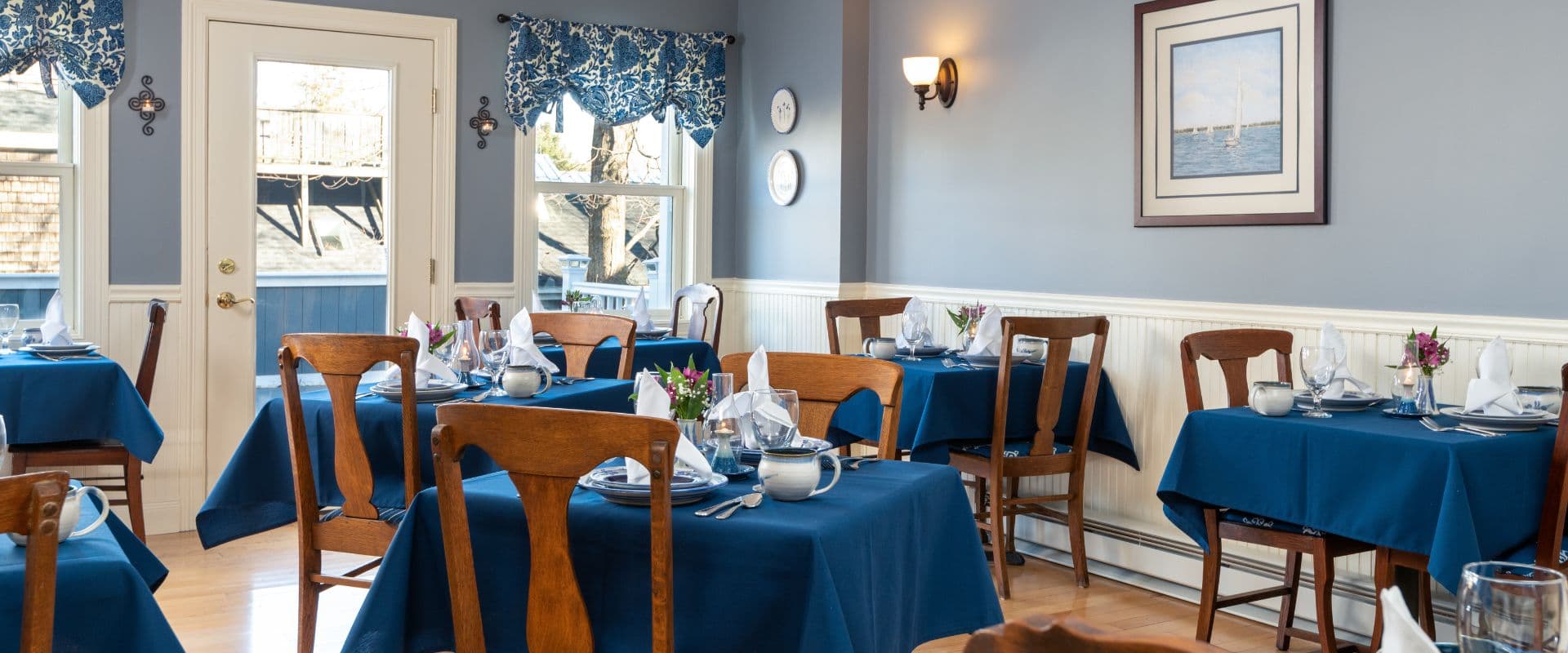 A cozy dining room with blue tablecloths and wooden chairs, featuring elegant table settings and coastal-themed decor.