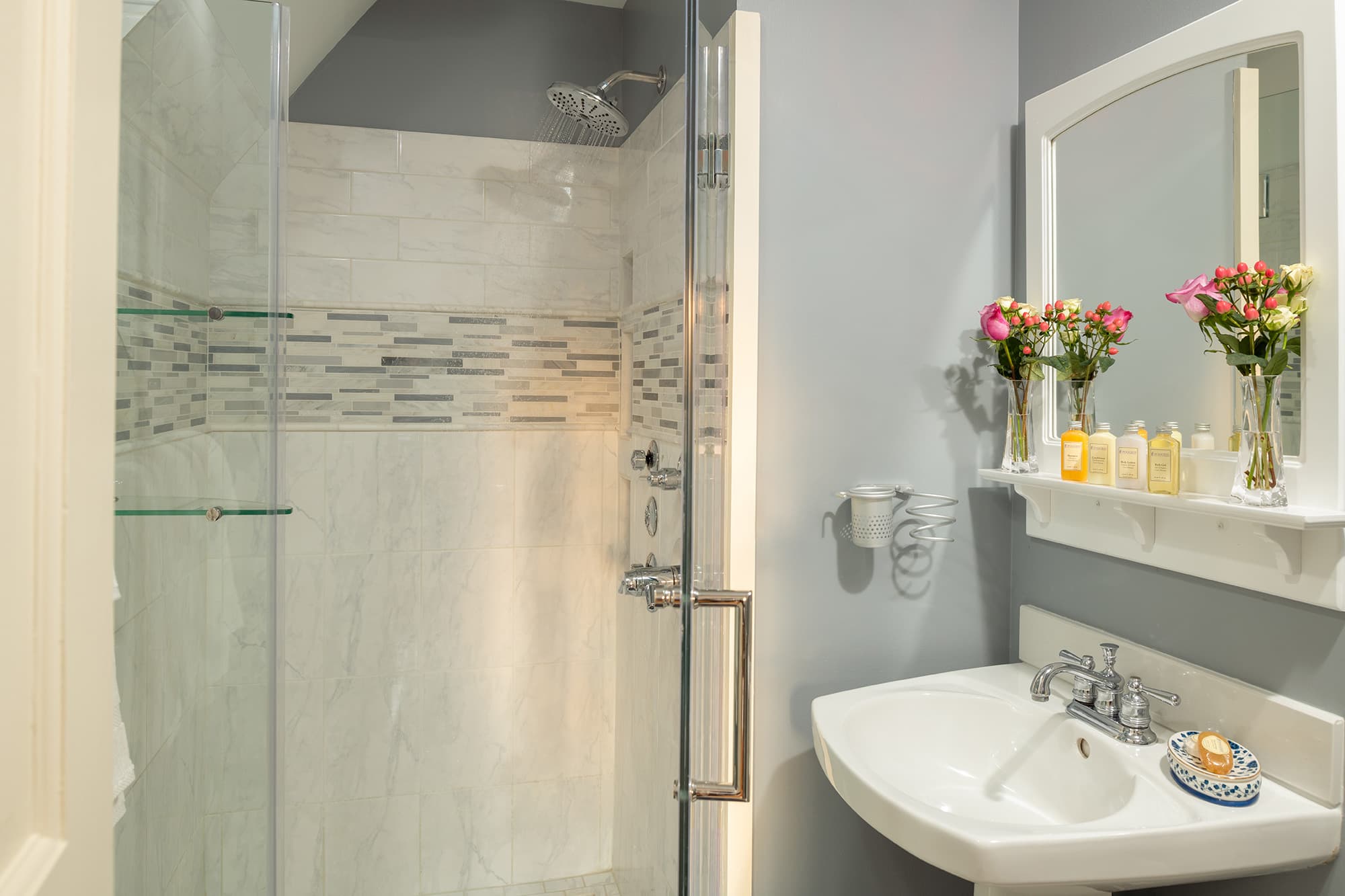 A bright, modern en-suite bathroom featuring a white pedestal sink, a glass-enclosed walk-in shower with a decorative tile border, and a white-framed mirror adorned with fresh pink roses.