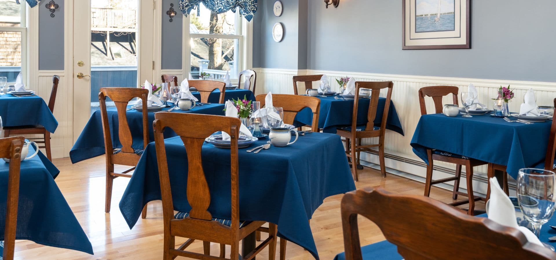 A cozy dining room featuring wooden tables with blue tablecloths, set with cutlery and small decorative flowers.