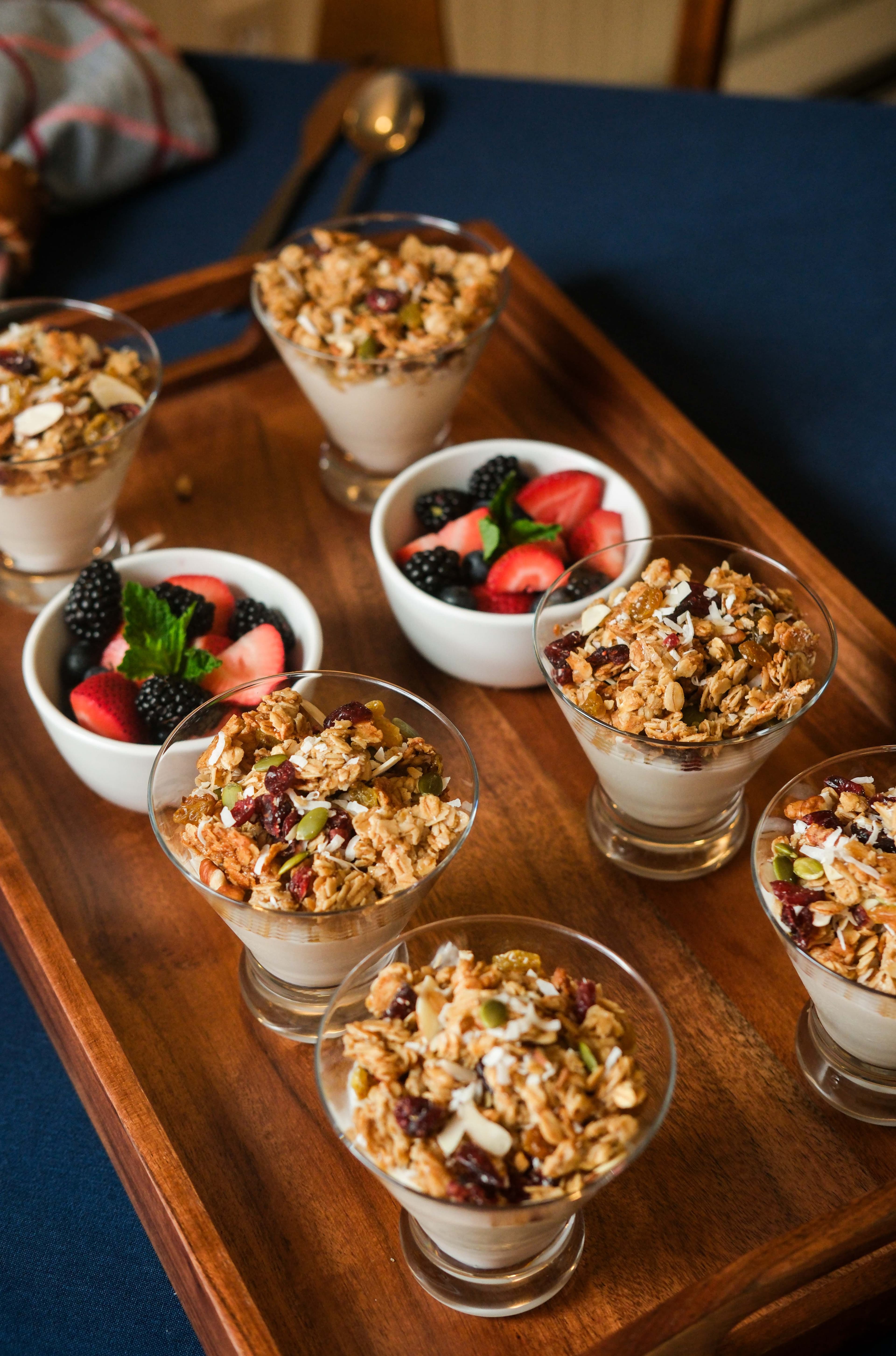A wooden tray holds several parfait glasses filled with granola and yogurt, alongside a bowl of mixed berries. A wooden tray holds several parfait glasses filled with granola and yogurt, alongside a bowl of mixed berries.