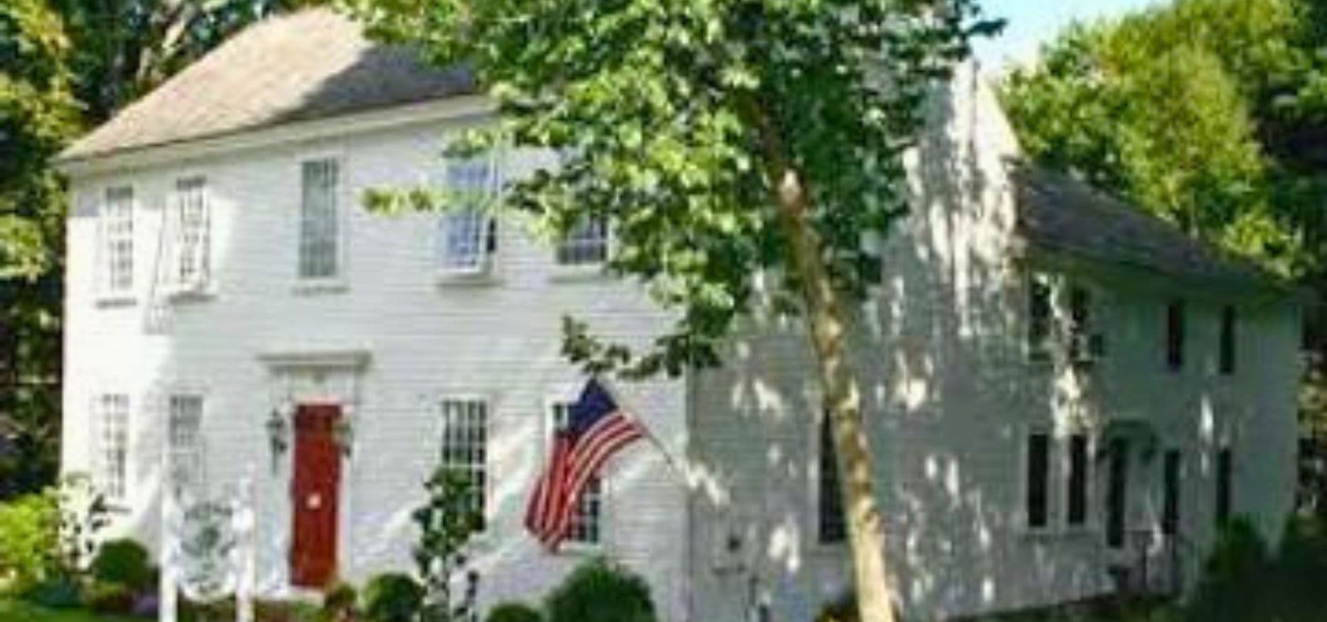 A white two-story house with a red door and an American flag, surrounded by greenery.