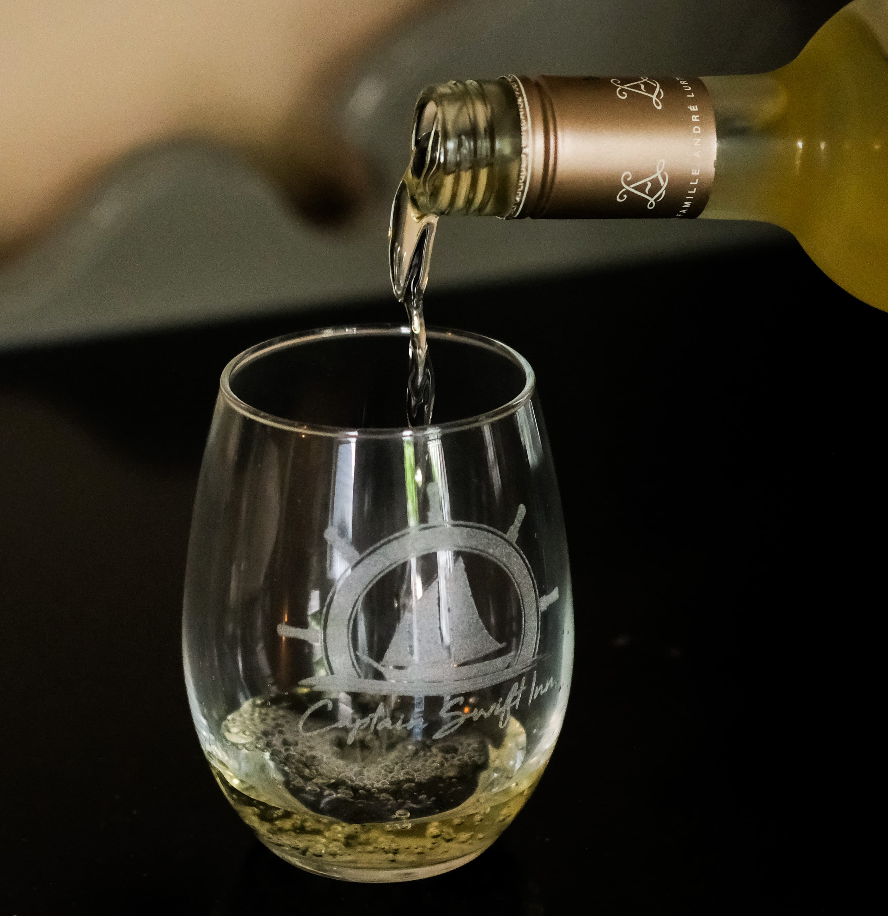 A bottle pours white wine into a branded glass.