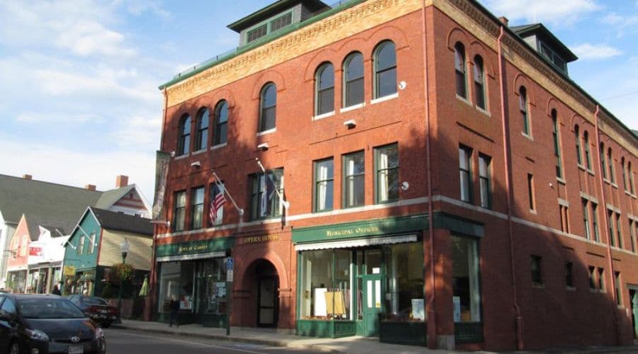 A red brick building with large windows, featuring flags and storefronts, set against a clear sky.