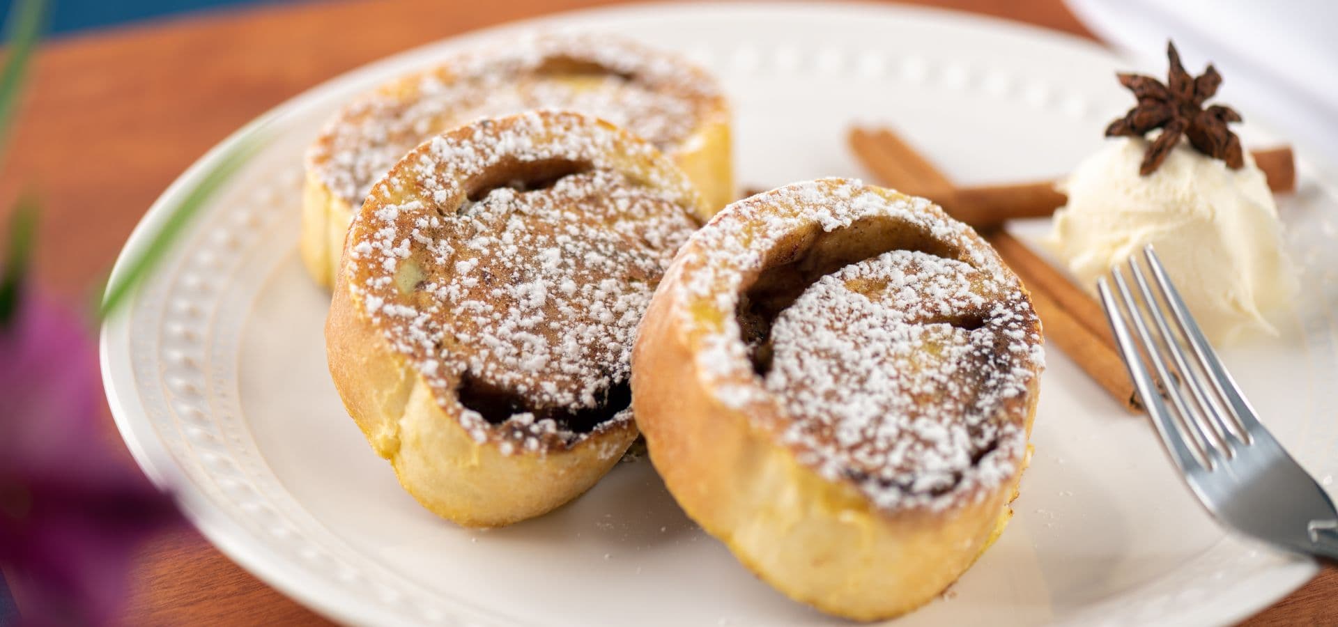 Three cinnamon rolls dusted with powdered sugar, served with a dollop of cream and a cinnamon stick on the side.