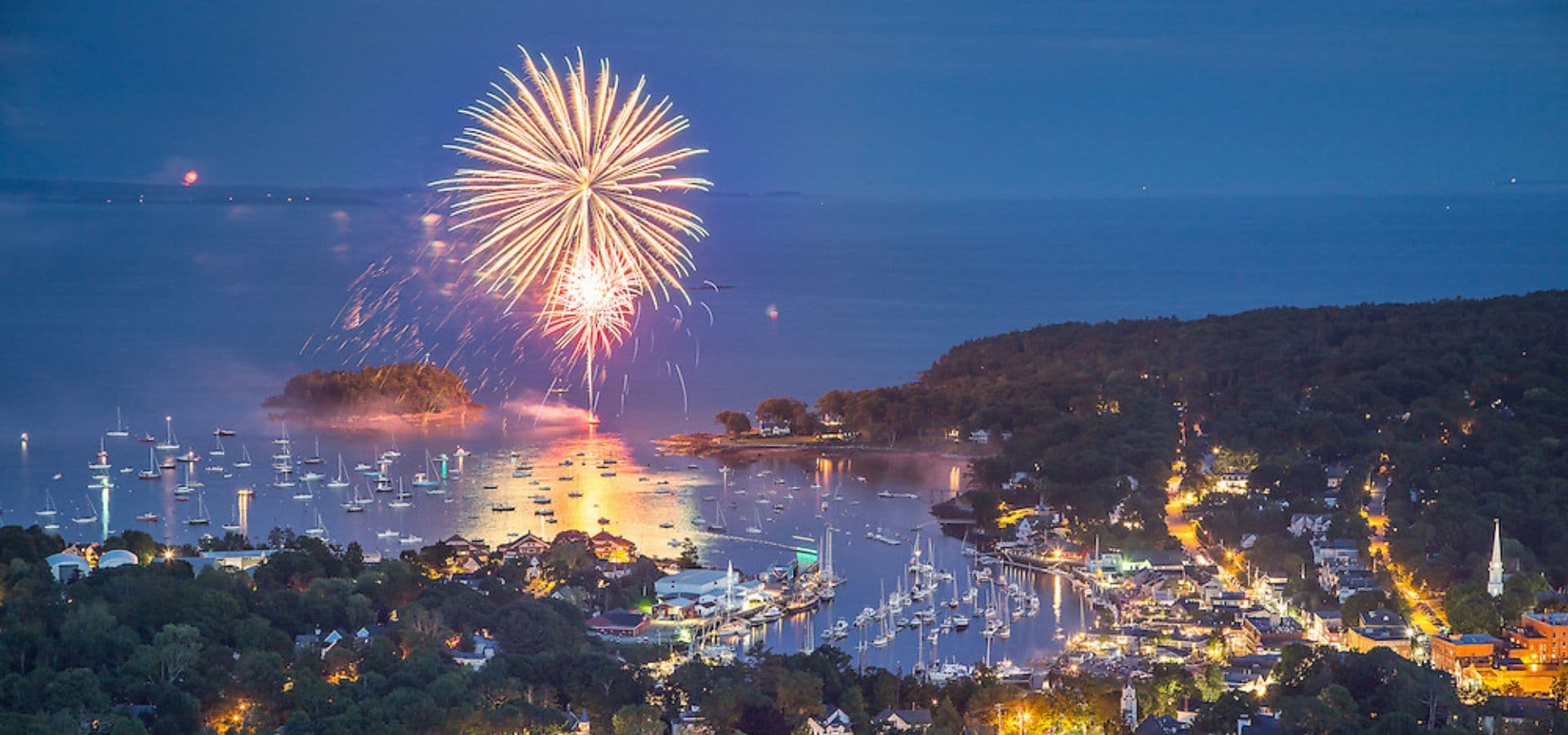 Fireworks illuminate the night sky over a harbor filled with boats and a coastal town.