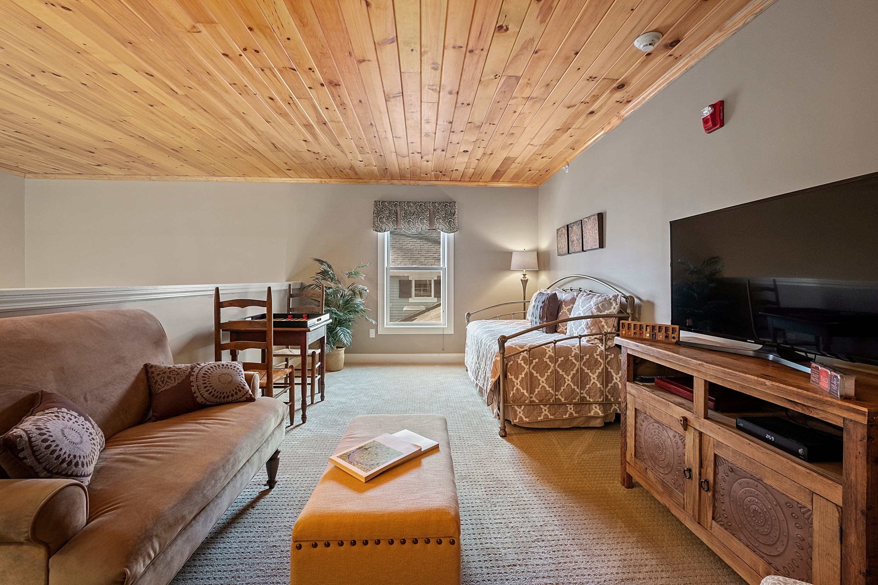 A cozy loft seating area with a natural wood plank ceiling, featuring a brown upholstered settee, a twin-size daybed, a game table with chairs, and a large flat-screen TV on a wooden console.