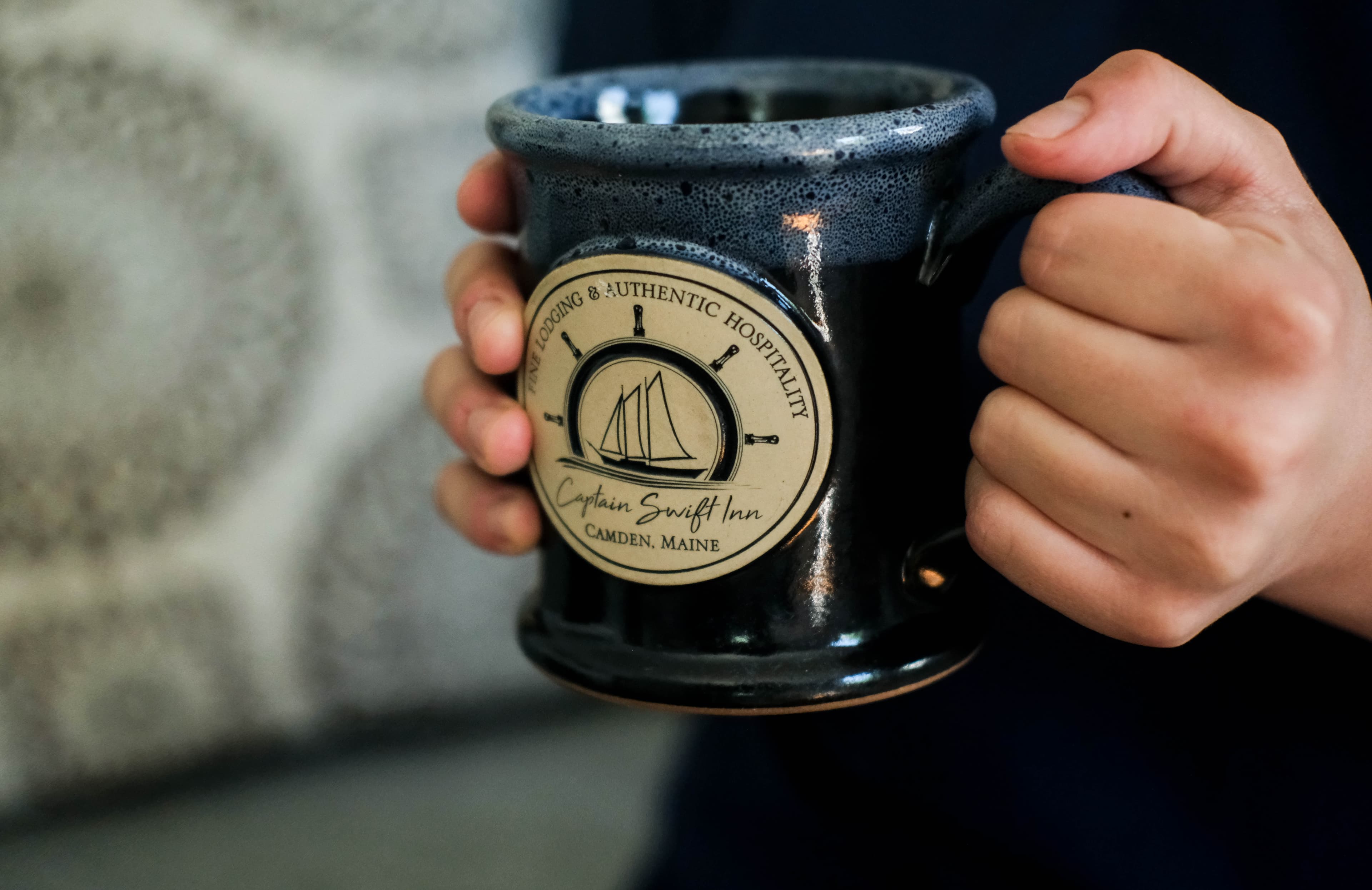 A person holding a blue ceramic mug with a logo from Captain Swift Inn in Camden, Maine.