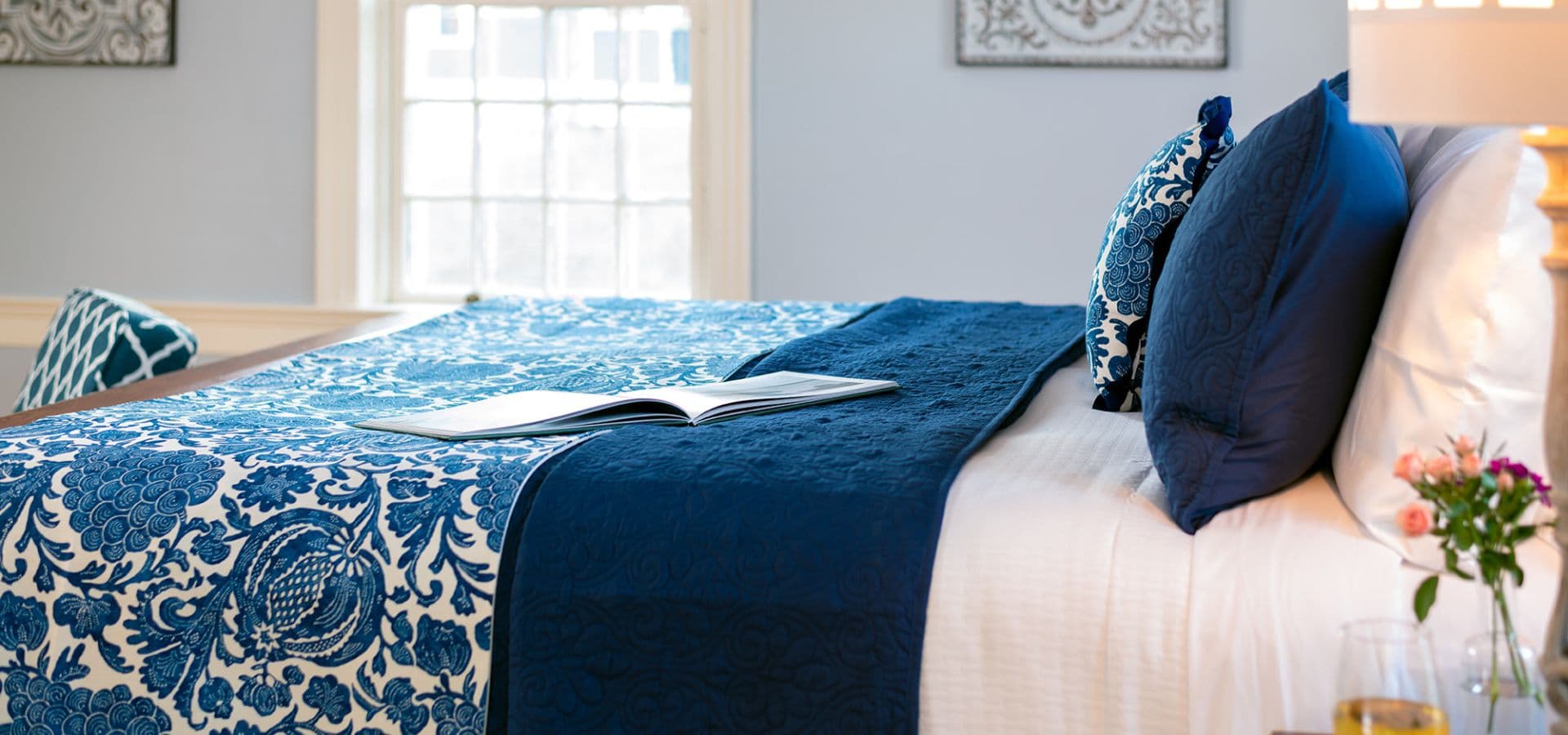 A neatly made bed with blue and white patterned bedding, a magazine, and decorative pillows beside a window.