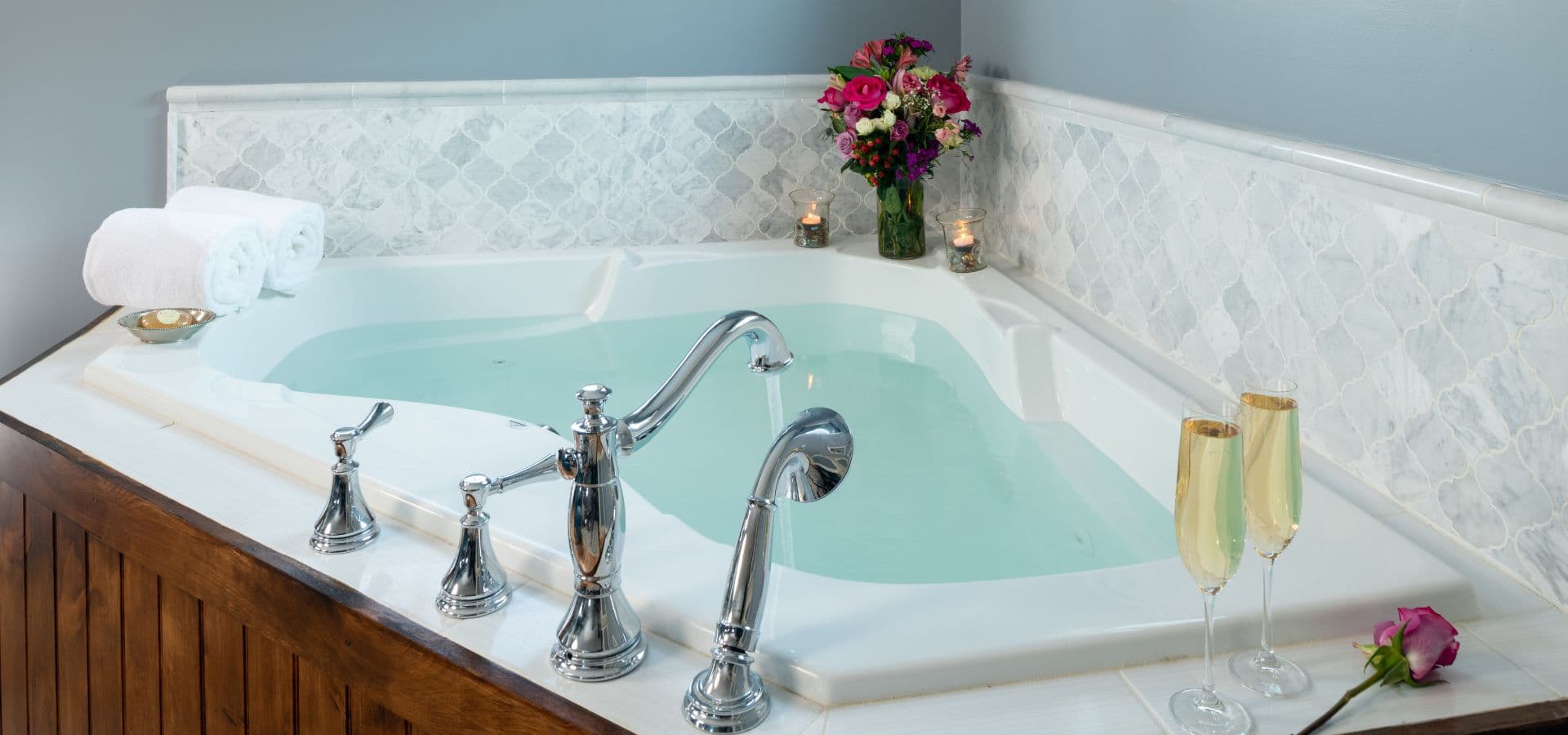 A luxurious jetted bathtub with champagne glasses and a bouquet of flowers nearby.