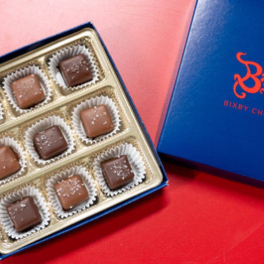 A box of assorted chocolates with a blue lid displays nine pieces, each in a paper cup.