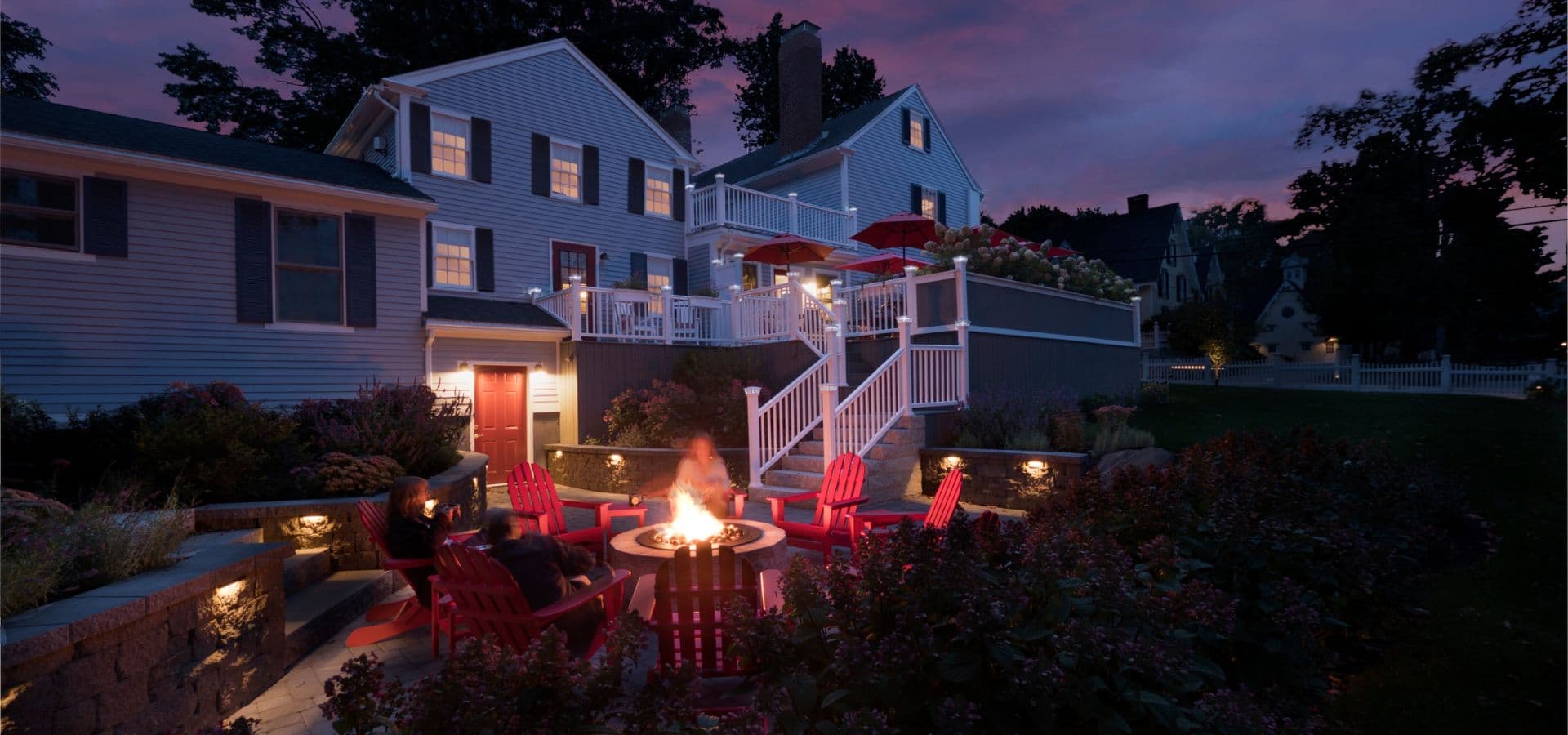 A cozy outdoor seating area with a fire pit, surrounded by colorful chairs, against the backdrop of a dusk sky and a two-story house.
