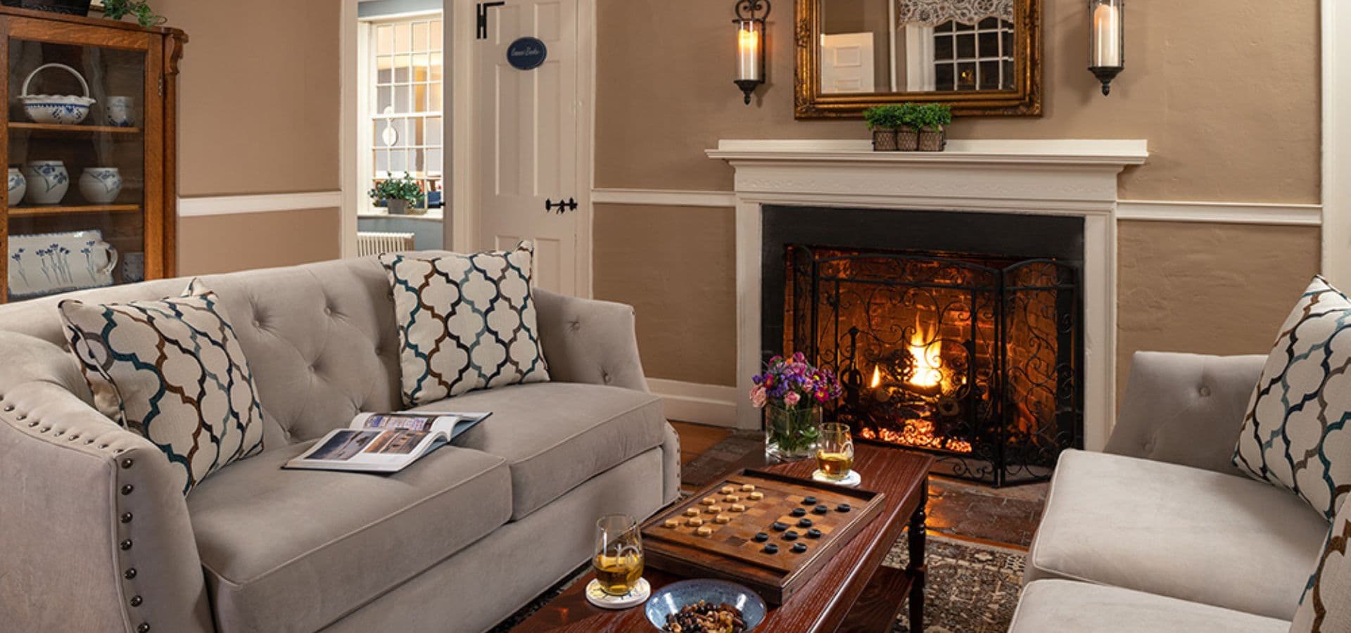 A cozy living room featuring a fireplace, comfortable sofas with patterned cushions, and a wooden coffee table with drinks and a game.