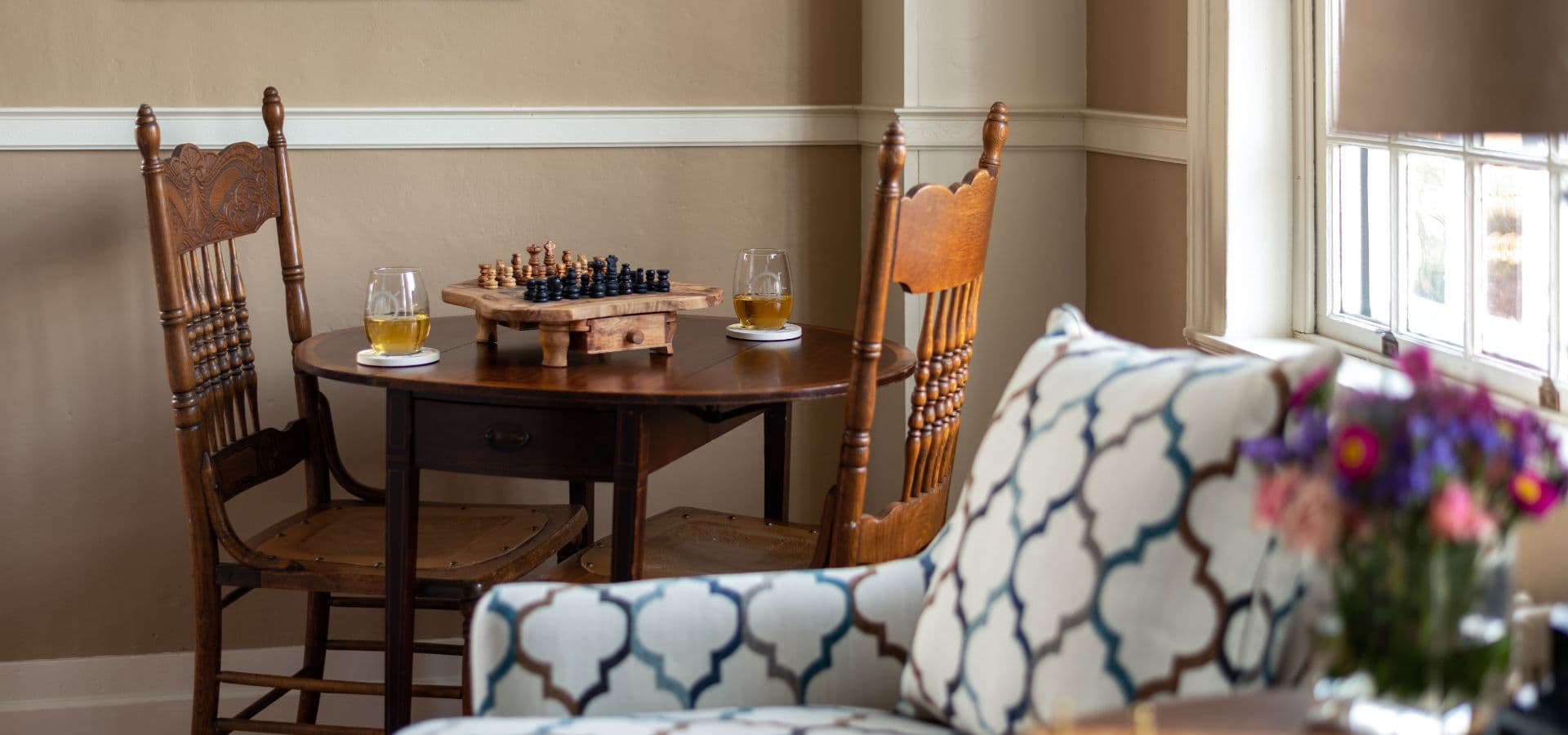 A cozy corner with a chess set on a small table, two chairs, and a stylish couch adorned with flowers.