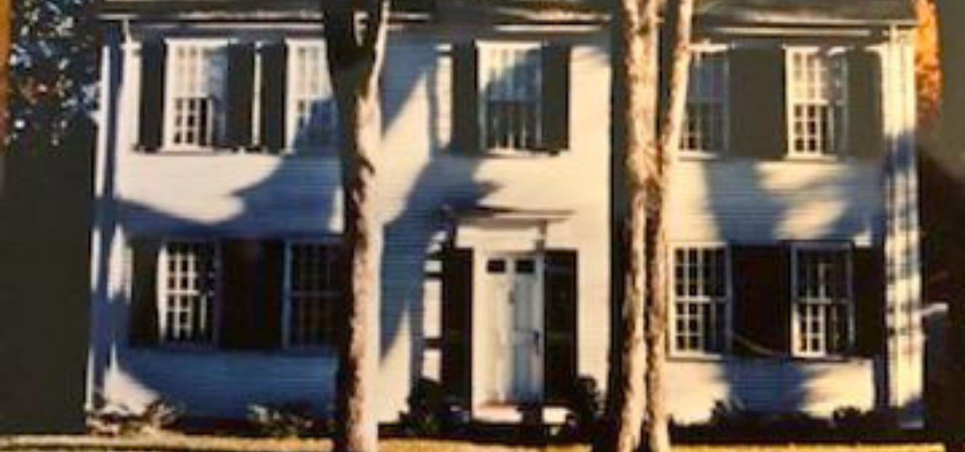 Two-story white house with black shutters and a front door, surrounded by trees.